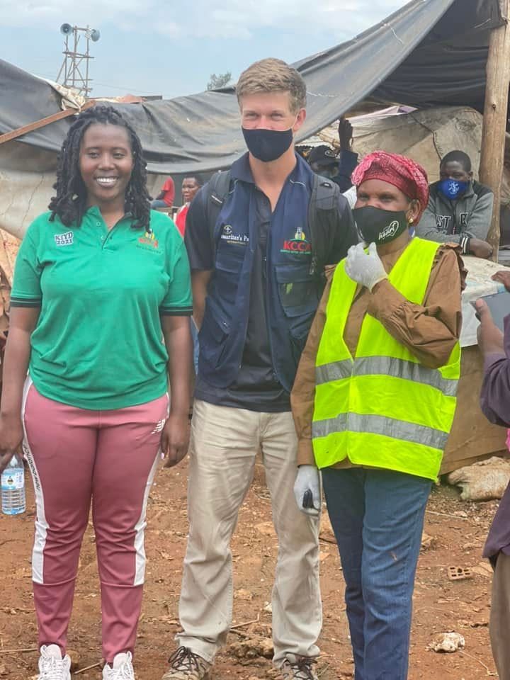 Three community workers, one in a green shirt and one in a high-visibility vest, stand together at a community site