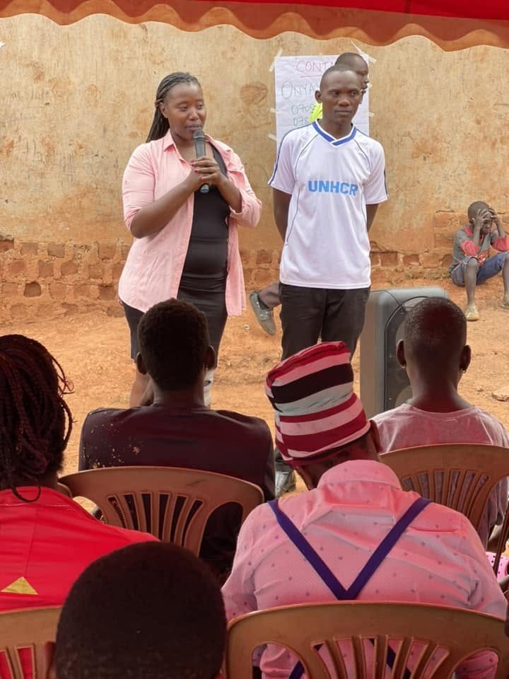 A woman speaks into a microphone to a seated community audience at an outdoor gathering