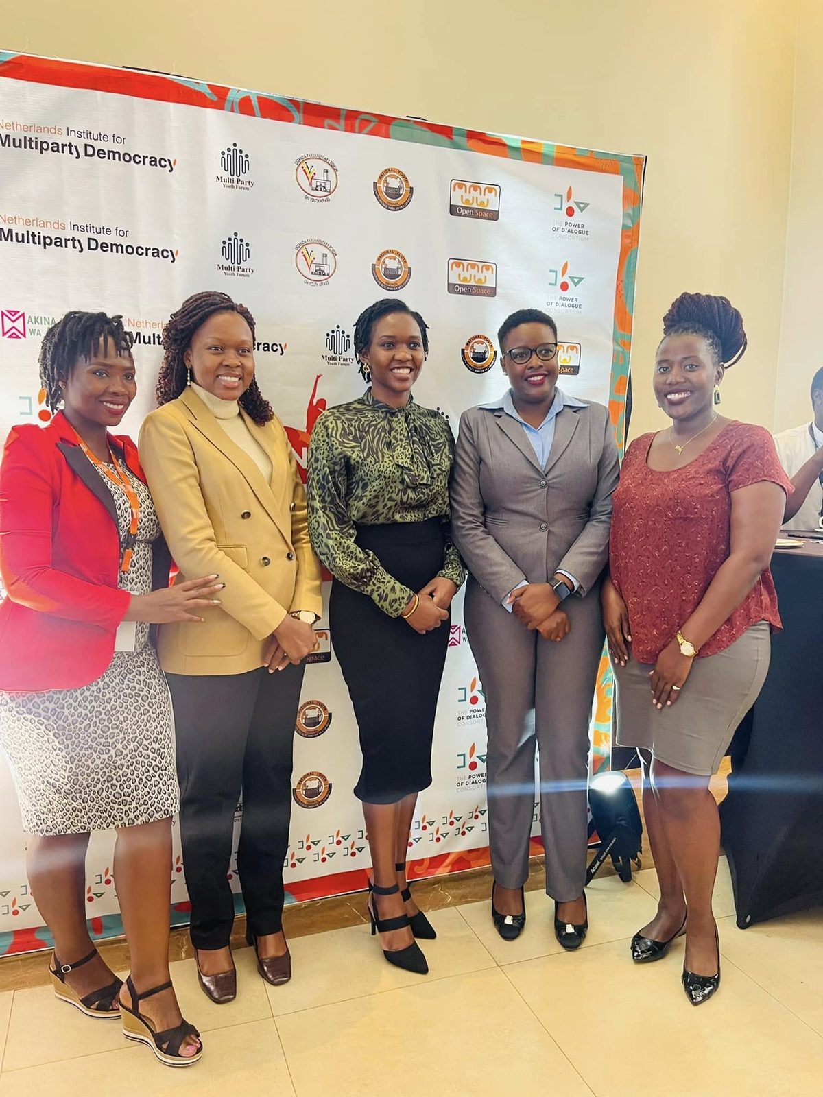 Five women in professional dress stand together in front of a partner backdrop at a policy event