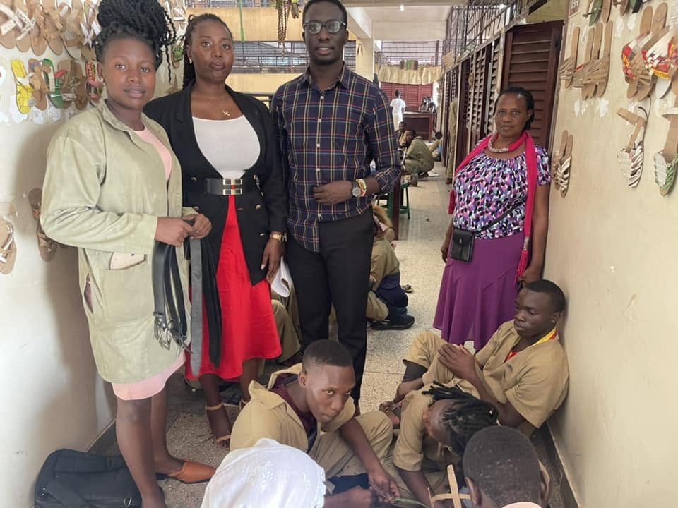 Trainees in workshop uniforms and visitors stand in a craft workshop lined with handmade sandals