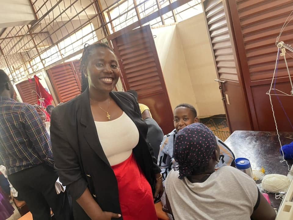A smiling woman in a black blazer stands in a knitting workshop while young women work at knitting machines
