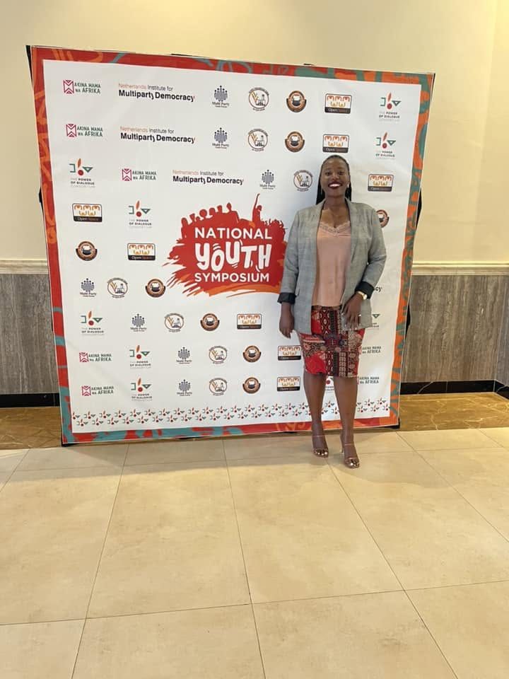 A woman stands in front of the National Youth Symposium backdrop featuring partner logos